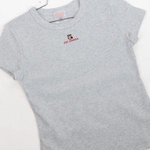 May include: A heather gray short-sleeve t-shirt featuring a small embroidered owl graphic centered on the chest above the red text "CHI OMEGA". The shirt has a crew neckline and is displayed on a metal hanger with a white brand label inside.