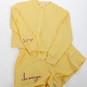 Può includere: Un completo due pezzi giallo brillante composto da un cardigan a maniche lunghe con bottoni e pantaloncini abbinati. La manica destra del cardigan e la parte inferiore della gamba sinistra dei pantaloncini presentano la scritta Chi Omega ricamata.
