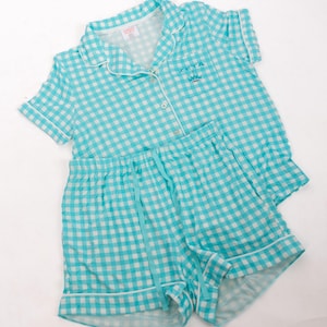 May include: A turquoise and white gingham pajama set consisting of a short-sleeved button-down shirt with a collar and matching shorts with a drawstring waist. The shirt features a small embroidered crown logo and the text ZETA on the pocket.