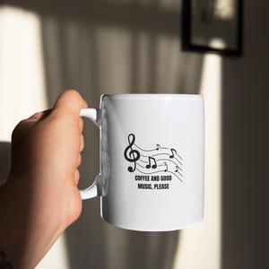 Puede incluir: Taza de cerámica blanca con un diseño de notas musicales negras y el texto "COFFEE AND GOOD MUSIC, PLEASE". La taza tiene un asa curva. El fondo es de color neutro.