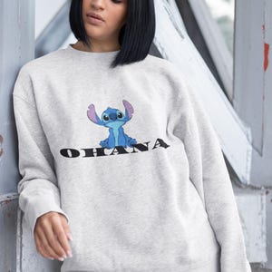 May include: A light gray sweatshirt featuring a blue cartoon character and the word "OHANA" in black. The sweatshirt has long sleeves and a crew neck. The design is centered on the front of the garment.