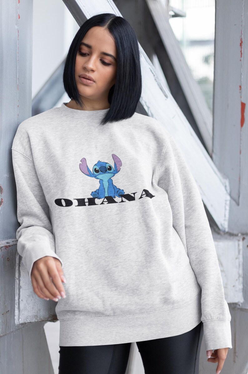 May include: A light gray sweatshirt featuring a blue cartoon character and the word "OHANA" in black. The sweatshirt has long sleeves and a crew neck. The design is centered on the front of the garment.