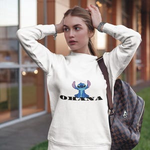 May include: White sweatshirt featuring a blue cartoon character and the word "OHANA" in black. The sweatshirt has long sleeves and a crew neck. The person is wearing blue jeans and a brown and black backpack.