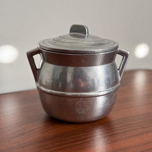 May include: A small, metallic, silver-colored lidded pot with two side handles. The pot features a rounded body with a horizontal ridge and a circular lid with a central handle. There are two small, circular stamped markings on the upper rim.