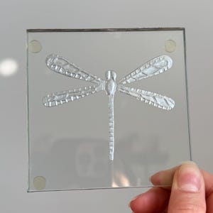 May include: A square, clear glass coaster featuring a centred, embossed silver dragonfly design. The coaster has four small, circular, translucent adhesive bumpers attached to the corners on the surface to provide elevation and grip.