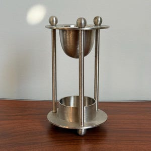 May include: A silver-toned metal stand featuring a circular base with three small spherical feet, three vertical support rods, and a top ring with three decorative spheres. A bowl-shaped vessel is suspended at the top, and a cylindrical base sits below.