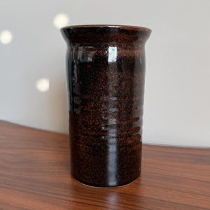 May include: A cylindrical ceramic vase with a dark brown, speckled glaze. The surface features subtle horizontal ridges along the body and a smooth, flared rim. The finish is glossy, reflecting light across the deep brown, earthy tones of the vessel.