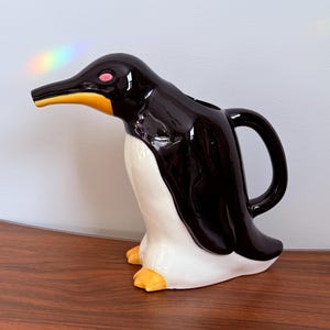 Op de afbeelding: Een keramische, pinguïnvormige kan met een glanzend zwarte body, witte buik en gele snavel en poten. De kan heeft een handvat en een tuit en is ontworpen om op een pinguïn te lijken. De kan is een decoratief item.