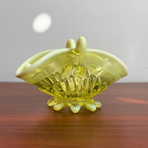 May include: A pale yellow glass basket featuring a scalloped base, a ribbed body with vertical fluting, and a smooth, curved rim. The piece includes a central arched handle that connects to the sides, showcasing a translucent finish.