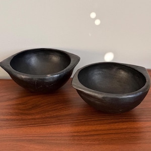 May include: Two matte black bowls with a rounded shape and distinct, pointed side handles. The bowls feature a smooth, dark finish and a deep, open interior. These vessels are crafted with a consistent dark tone and a simple, functional design.