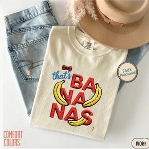 May include: An ivory t-shirt featuring embroidered text that reads that's BANANAS with three yellow banana illustrations and a small red and black plaid bow. The shirt is paired with light blue denim jeans, a tan wide-brimmed hat, and a silver necklace.