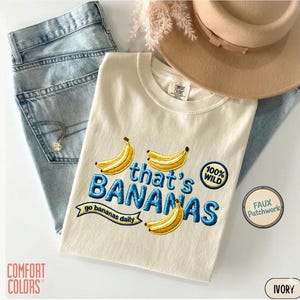 May include: An ivory t-shirt featuring embroidered yellow bananas and blue text that reads that's BANANAS, go bananas daily, and 100% WILD. The shirt is displayed with light blue denim jeans, a tan wide-brimmed hat, and a silver chain necklace.