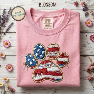 May include: A light pink t-shirt featuring an embroidered paw print design composed of faux patchwork patterns in red, white, and blue. The design includes stars and floral motifs. The inner neck label reads Comfort Colors with size L.