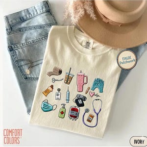 Può includere: Una t-shirt color avorio con icone mediche ricamate tra cui uno stetoscopio, una siringa, una mascherina, una sacca di sangue e un disinfettante. È abbinata a jeans azzurri, un cappello beige a tesa larga e una collana d'argento.