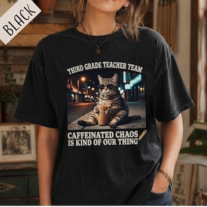 Puede incluir: Una camiseta negra de manga corta con un gráfico de un gato atigrado con gafas de sol sosteniendo un café helado en una calle. El texto dice THIRD GRADE TEACHER TEAM y CAFFEINATED CHAOS IS KIND OF OUR THING con un icono de un lápiz.