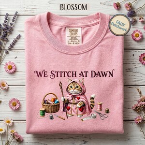 Könnte beinhalten: Ein hellrosa T-Shirt mit einer Grafik einer getigerten Katze, die einen rosa Kapuzenumhang trägt. Der Text "WE STITCH AT DAWN" steht über der Katze. Die Grafik zeigt einen Nähkorb, Garnrollen, eine Nadel und eine Schere.