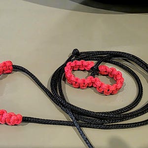 May include: A black cord lead featuring two sections of bright pink macrame knotting. The lead includes a large pink knotted handle loop on one end and two smaller pink knotted loops positioned along the length of the black cord.