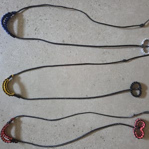 May include: Three handmade lanyards with black cords. The lanyards have decorative elements in blue, yellow, and red. Each lanyard has a loop at the end. The background is a light gray surface.