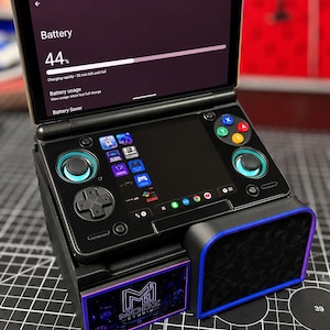 以下が含まれることがあります： デュアルスクリーンを備えた黒い携帯型ゲーム機。上画面にはバッテリー残量44%が表示され、下画面にはゲームアイコンが並んでいます。十字キー、2つのアナログスティック、4つのアクションボタンが搭載されています。