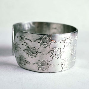 May include: A silver metal bracelet with a repeating pattern of bees embossed on the surface.