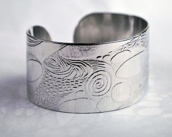 Puede incluir: Un brazalete de metal plateado con un intrincado diseño en relieve de patrones en espiral y formas redondeadas.