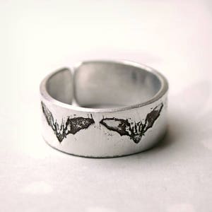 Puede incluir: Anillo plateado con superficie texturizada y diseño de murciélago. El anillo presenta dos siluetas de murciélago con detalles de alas. El anillo es de estilo banda ancha, adecuado para el uso diario. Diámetro aproximado de 2 cm.