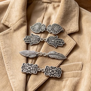 May include: A tan corduroy blazer featuring four distinct silver-toned metal clasps attached to the front. The clasps include designs of ornate hearts, hamsa hands, detailed feathers, and rectangular floral patterns, all with an antique finish.