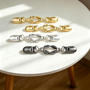 Può includere: Quattro fermagli metallici per abiti in oro, argento e nero disposti su una superficie bianca. Ogni fermaglio presenta un design a nodo decorativo al centro e una chiusura a ciascuna estremità. I fermagli sono progettati per tenere insieme il tessuto.