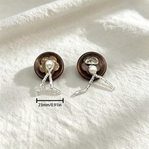 May include: Two circular dark brown buttons, each measuring 0.91 inches in diameter. Each button features a central white pearl bead, a small metallic flower or stone accent, and dangling clear glass seed beads arranged in a V-shape pattern.
