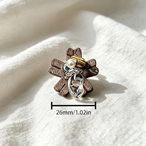 May include: A flower-shaped brooch made of dark brown wood with a visible grain, adorned with a white pearl, a gold-toned ribbed bead, and a clear glass accent. The brooch measures 26mm or 1.02 inches in width as indicated by the text.