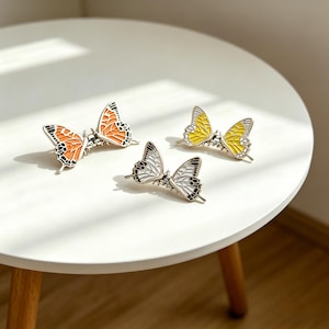 Puede incluir: Cuatro pinzas para el pelo con forma de mariposa en naranja, amarillo y blanco con detalles en negro. Las pinzas metálicas están dispuestas sobre una superficie blanca. Las alas de mariposa presentan patrones detallados.