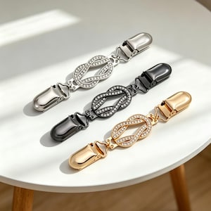 Könnte beinhalten: Drei dekorative Pulloverclips in Silber, Schwarz und Gold. Jeder Clip hat ein Knotendesign, das mit kleinen, funkelnden Strasssteinen verziert ist. Die Clips haben ein klassisches Design mit einem Scharnierverschluss an jedem Ende.