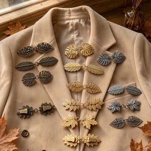 May include: A collection of metal cardigan clips in various leaf and botanical shapes, including gold, silver, and bronze tones. These fasteners feature intricate filigree, textured veins, and solid metal designs, arranged on a beige corduroy blazer.
