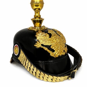 Op de afbeelding: Een zwarte militaire helm met gouden details. De helm heeft een gouden adelaarsinsigne, een decoratieve gouden band en een gouden top. Deze historische of ceremoniële helm is waarschijnlijk van militaire oorsprong.
