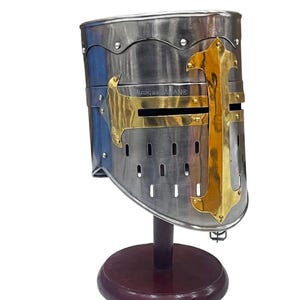 Puede incluir: Casco medieval plateado con un diseño de cruz dorada en la parte delantera. El casco, con construcción remachada, se exhibe sobre un soporte de madera marrón oscuro. El texto "Musigaa JALAN®" es visible en el lateral. Objeto histórico.