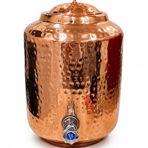 May include: A hammered copper water cooler with a lid and a spigot. The copper cooler has a textured surface and a small, round lid handle. The spigot has a blue handle. The cooler is approximately 14 inches tall.