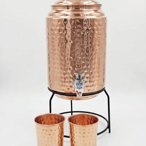 May include: A copper water dispenser with a hammered texture, a spigot, and a lid, set on a black metal stand. Two matching copper cups sit in front. The dispenser is designed to hold water and is a decorative item.