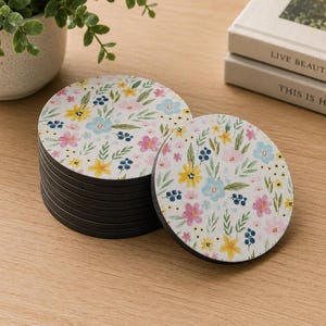 May include: A stack of round drink coasters with a white surface featuring a colorful floral pattern of yellow, pink, and light blue flowers with green leaves and dark blue berries. The coasters have a solid black base and a smooth, flat top surface.