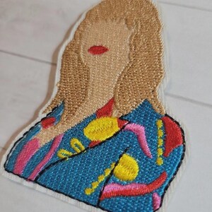 May include: Embroidered patch featuring a stylized portrait of a person with blonde hair and red lips. The person is wearing a blue jacket with yellow, pink, and red abstract designs. The patch has a white border and black stitching.