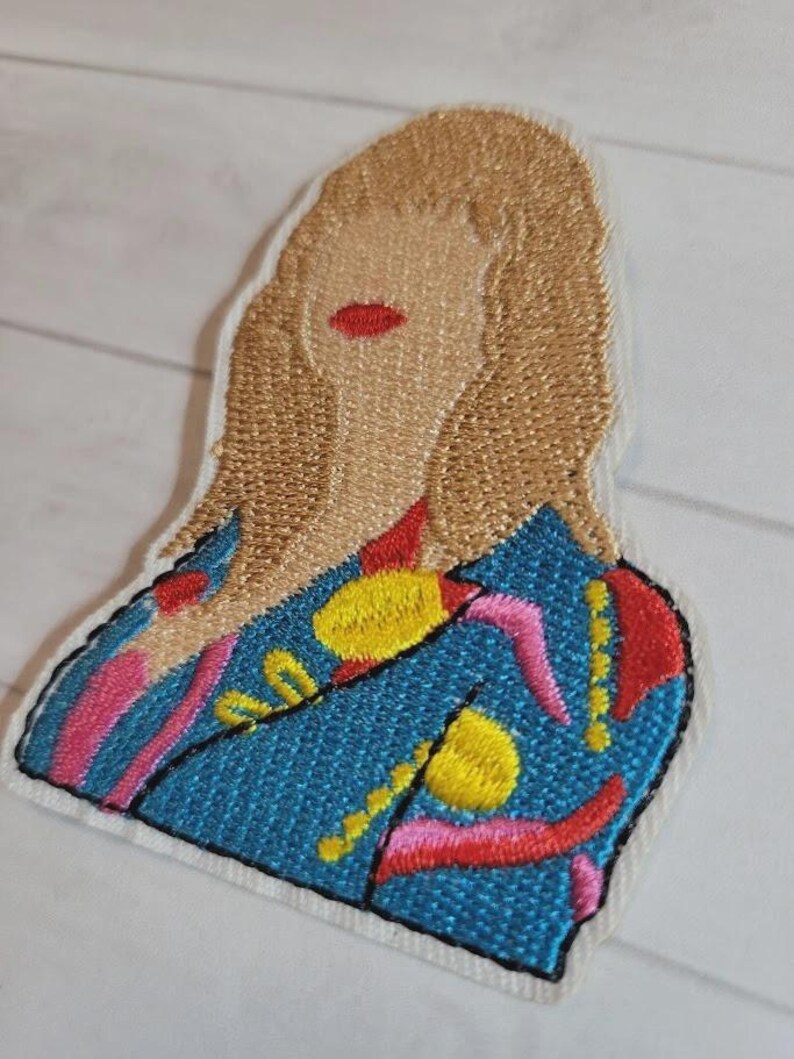 May include: Embroidered patch featuring a stylized portrait of a person with blonde hair and red lips. The person is wearing a blue jacket with yellow, pink, and red abstract designs. The patch has a white border and black stitching.