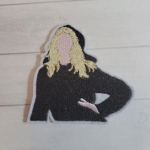 May include: Embroidered patch featuring a stylized figure with blonde hair, a pink face, and a black top. The patch has a white border and is likely designed for application on clothing or accessories. The design is simple and modern.