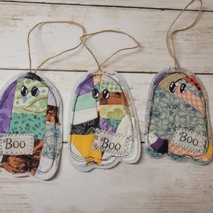 May include: Three ghost-shaped fabric ornaments featuring patchwork designs in various colors and patterns. Each ghost has stitched black eyes and a white rectangular patch at the bottom with the word Boo printed in black text, attached to a twine loop.