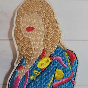 May include: Embroidered patch featuring a stylized portrait of a person with blonde hair and red lips. The person is wearing a blue jacket with yellow, pink, and red embroidered details. The patch has a white border.