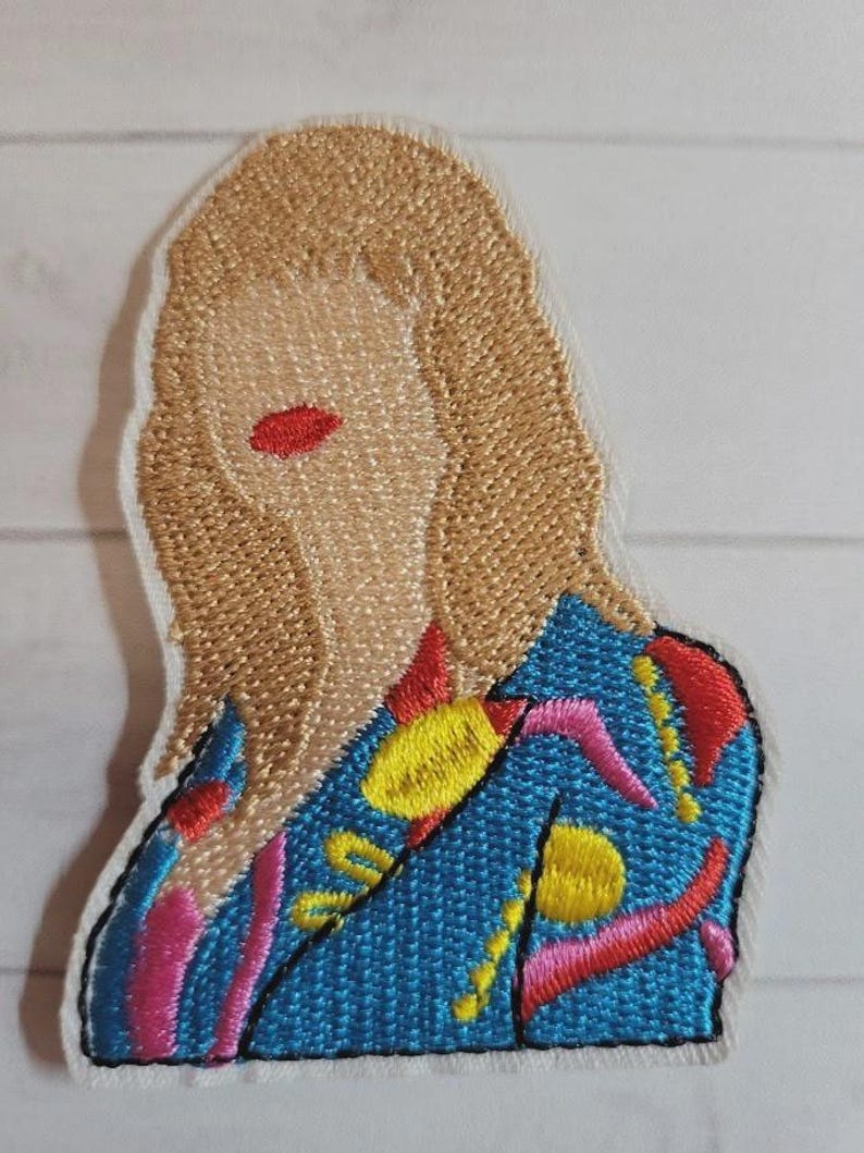 May include: Embroidered patch featuring a stylized portrait of a person with blonde hair and red lips. The person is wearing a blue jacket with yellow, pink, and red embroidered details. The patch has a white border.