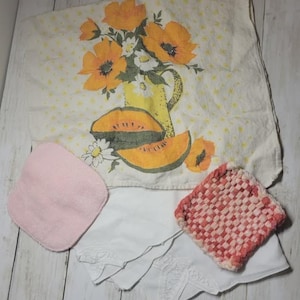 May include: A collection of vintage kitchen linens, including a floral-printed tea towel with orange poppies and a yellow pitcher, a pink washcloth, a white embroidered napkin, and a red and white woven potholder. The items are arranged on a light wood surface.
