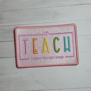May include: A rectangular pink embroidered patch with the word "TEACH" in colorful letters and the phrase "THEM TO BE KIND" below. A small pink heart is above the word "TEACH".