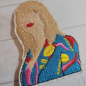 May include: Embroidered patch featuring a stylized figure with blonde hair and red lips. The figure wears a blue jacket with colorful abstract designs in pink, yellow, and red. The patch has a white border and is likely for clothing or accessories.