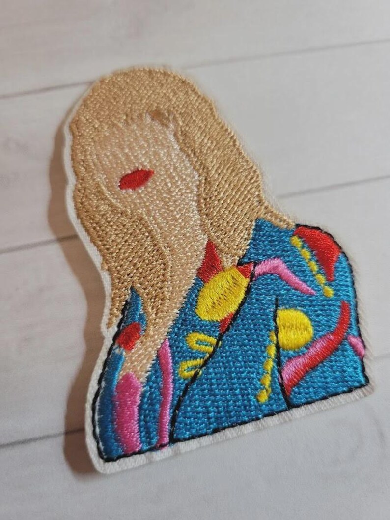 May include: Embroidered patch featuring a stylized figure with blonde hair and red lips. The figure wears a blue jacket with colorful abstract designs in pink, yellow, and red. The patch has a white border and is likely for clothing or accessories.
