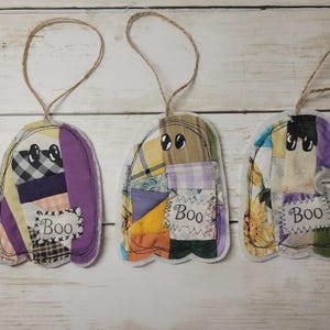 May include: Three ghost-shaped fabric ornaments featuring patchwork designs in various colors and patterns. Each ornament has two black eyes and a white rectangular patch with the word Boo stitched in black thread. Each includes a jute twine loop.