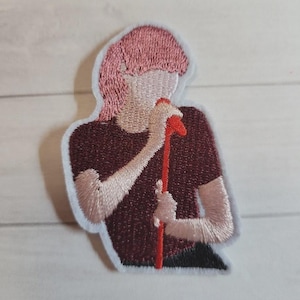 May include: Embroidered patch featuring a figure with pink hair holding a red microphone. The figure is wearing a maroon shirt and has a white outline. The patch is likely for clothing or accessories.